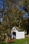 Small white chapel under a tree with a red-brown bench in front of it