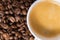 Small white coffee cup with espresso crema on roasted beans background. Close-up. Selective focus