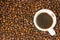 Small white cup of brewed coffee with steam above it standing on a background of scattered brown roasted coffee beans