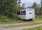 Small white trailer house with blue sign office, abandoned caravan in rural landscape near footpath, green grass and trees