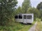 Small white trailer house with blue sign office, abandoned caravan in rural landscape near footpath, green grass and trees