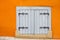 A small white window is closed by wooden shutters with decorative hinges on a bright orange wall