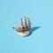 Small wooden home decoration pirate ship with white sails on donut with white glaze and colorful crumbs. Blue pastel ackground.