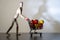 Small wooden mannequin pushing a mini supermarket cart with colorful peppers