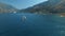 Small yacht sails along the Bay of Kotor, behind it there are beautiful mountains and cozy little town