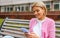 Smart blonde young woman wearing blue jeans, white t-shirt and pink jacket, smiling and sending messages on smart phone. Pretty