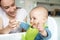 Smiling 8 Month Old Baby Boy At Home In High Chair Being Fed Solid Food By Mother With Spoon