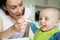 Smiling 8 month Old Baby Boy At Home In High Chair Being Fed Solid Food By Mother With Spoon