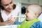 Smiling 8 month Old Baby Boy At Home In High Chair Being Fed Solid Food By Mother With Spoon