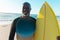 Smiling african american bald senior man with yellow surfboard standing against sea and clear sky