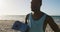 Smiling african american man holding yoga mat, standing on beach by the sea