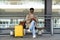 Smiling Afro-American traveler man with yellow suitcase sitting on bench in airport terminal or railway station, using mobile