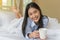Smiling Asian young woman lying holding a cup of coffee on bed