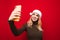 Smiling attractive girl in sledge claus hat makes selfie on red background, looks at smartphone and smiles. Attractive lady in a