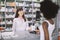 Smiling attractive young red head Caucasian pharmacist offering medicine to young African female patient, paying for the