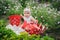 Smiling baby is sitting on the green grass and flowers with dotted red bucket and watering can.