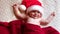 Smiling Baby Wearing Red Santa Claus Hat Celebrating Christmas. Cute Newborn Baby In Christmas Hat Lying On Knit Blanket