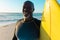 Smiling bald african american senior man with yellow surfboard standing against sea and clear sky