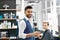 Smiling barber standing near young client sitting near mirror.