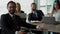 Smiling bearded businessman in black suit is sitting by the desk. Colleagues are woking on the background.