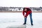 Smiling bearded man in a red jacket and jeans pierces the ice on the frozen river with a crowbar