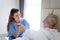 Smiling beautiful caring female doctor holding hand of male senior patient who lying in hospital bed. Nurse takes care elderly man