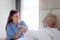 Smiling beautiful caring female doctor holding hand of male senior patient who lying in hospital bed. Nurse takes care elderly man