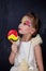 Smiling beautiful little girl with lollipop in white dress red lips with painted face at dark background. Children love candy