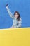 Smiling beautiful woman lean on yellow wall looking camera while holding a mobile phone up in the air