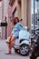 Smiling beautiful woman with long brown hair dressed in trendy clothes posing with white retro Italian scooter on the