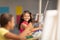 Smiling biracial elementary schoolgirl looking at female classmate during drawing class in school