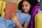 Smiling biracial elementary schoolgirl reading book while lying on couch in school