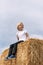 Smiling blond boy in white T-shirt sits in the hay and looks into the distance. Portrait of child on haystack