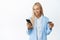 Smiling blond corporate woman enter info on mobile phone, holding credit card, wearing suit, standing over white