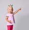 Smiling blonde baby girl in pink girlish casual clothing and princess crown stands pointing with finger at upper corner