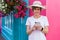 Smiling Blonde Tourist: Casual, Hat-Clad, Texting in Vibrant Ireland with Colorful Houses and Floral Decor