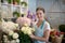 Smiling brown-haired female in apron standing in front of flower stand, touching roses