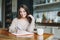 Smiling brunette asian young woman in casual doing notes in daily book with cup of tea on kitchen at the home