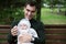 Smiling brunette dad in reen jacket sits on bench with baby in his arms in Park