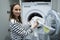 Smiling brunette, a decent housewife, stands in the laundry room and loads the washing machine with dirty clothes