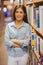 Smiling brunette student standing next to bookshelves with arms crossed