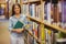 Smiling brunette student standing next to bookshelves while holding books