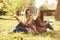 Smiling brunette twin girls sitting back to back on the grass and looking at each other, legs slightly bent in knees