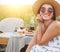 Smiling at camera young Sincerely  Woman in sunglasses and straw hat dressed light summer dress sitting in rattan chair on the