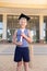 Smiling caucasian elementary schoolboy with mortarboard and degree standing against school building