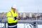 A smiling caucasian foreman in a white helmet and a yellow vest holds clipboard, stands on the roof