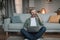 Smiling caucasian retired male with beard sits on floor with laptop, calls by phone in living room