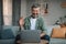 Smiling caucasian retired man with beard waves hand, looks at laptop in living room interior