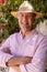 Smiling caucasian senior man wearing hat and eyeglasses with arms crossed standing against plants