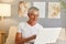 Smiling charming grey-haired mature man wearing casual white T-shirt and glasses sitting on sofa at home interior studying how to